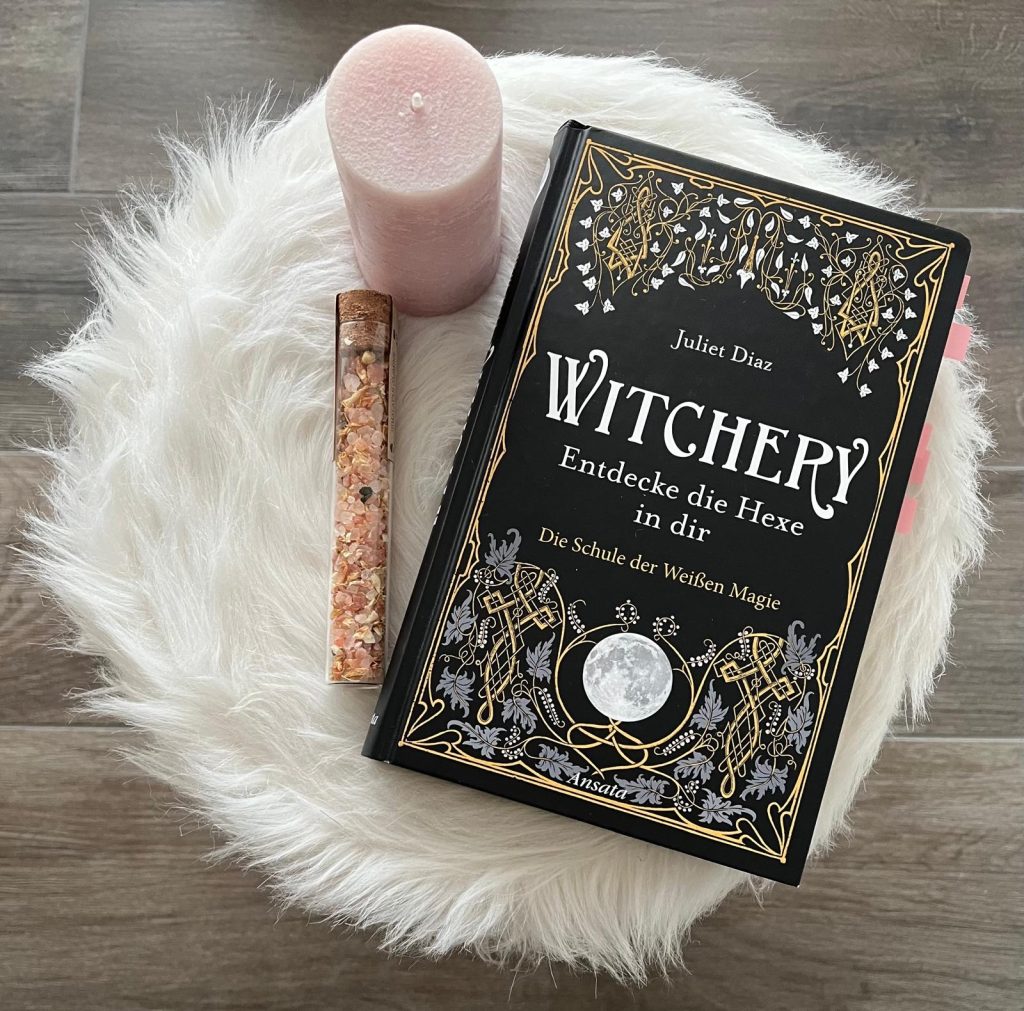 Das Sachbuch "Witchery: Entdecke die Hexe in dir" auf weissem Fell mt einer rosa Kerze und pinken Steinchen.