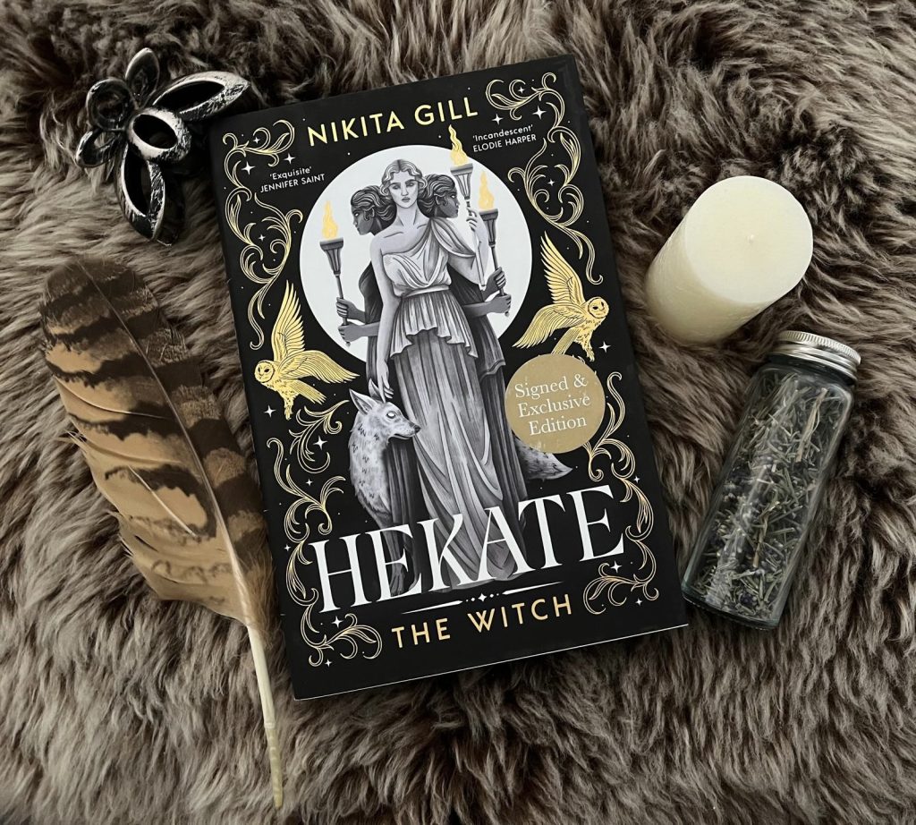Der Roman "Hekate: The Witch" von Nikita Gill auf dunklem Fell mit einer Feder, einer Kerze, Lavendelblüten und silberner Dekoration.