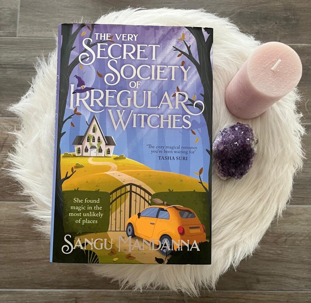 The book “The very secret society of irregular witches” by Sangu Mandanna on white fur next to a pink candle and a cristal.