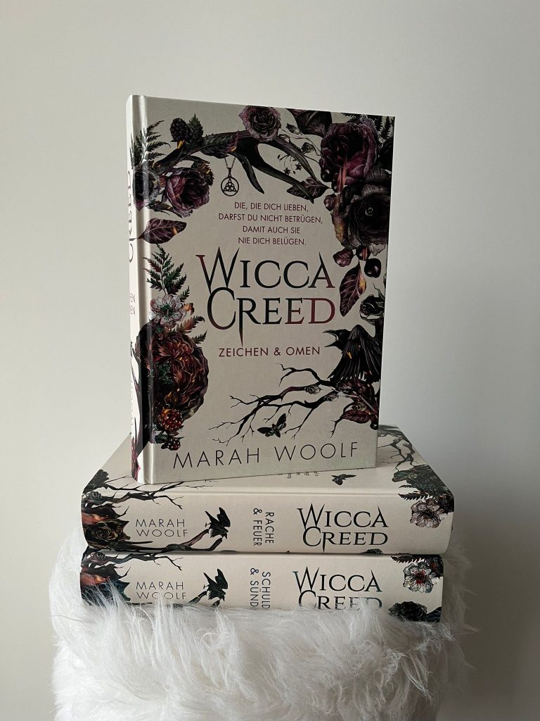 Three volumes of the "Wicca Creed" series by Marah Woolf, decoratively stacked against a light-colored wall with floral and dark cover designs, with the first book, "Zeichen und Omen", placed on top.